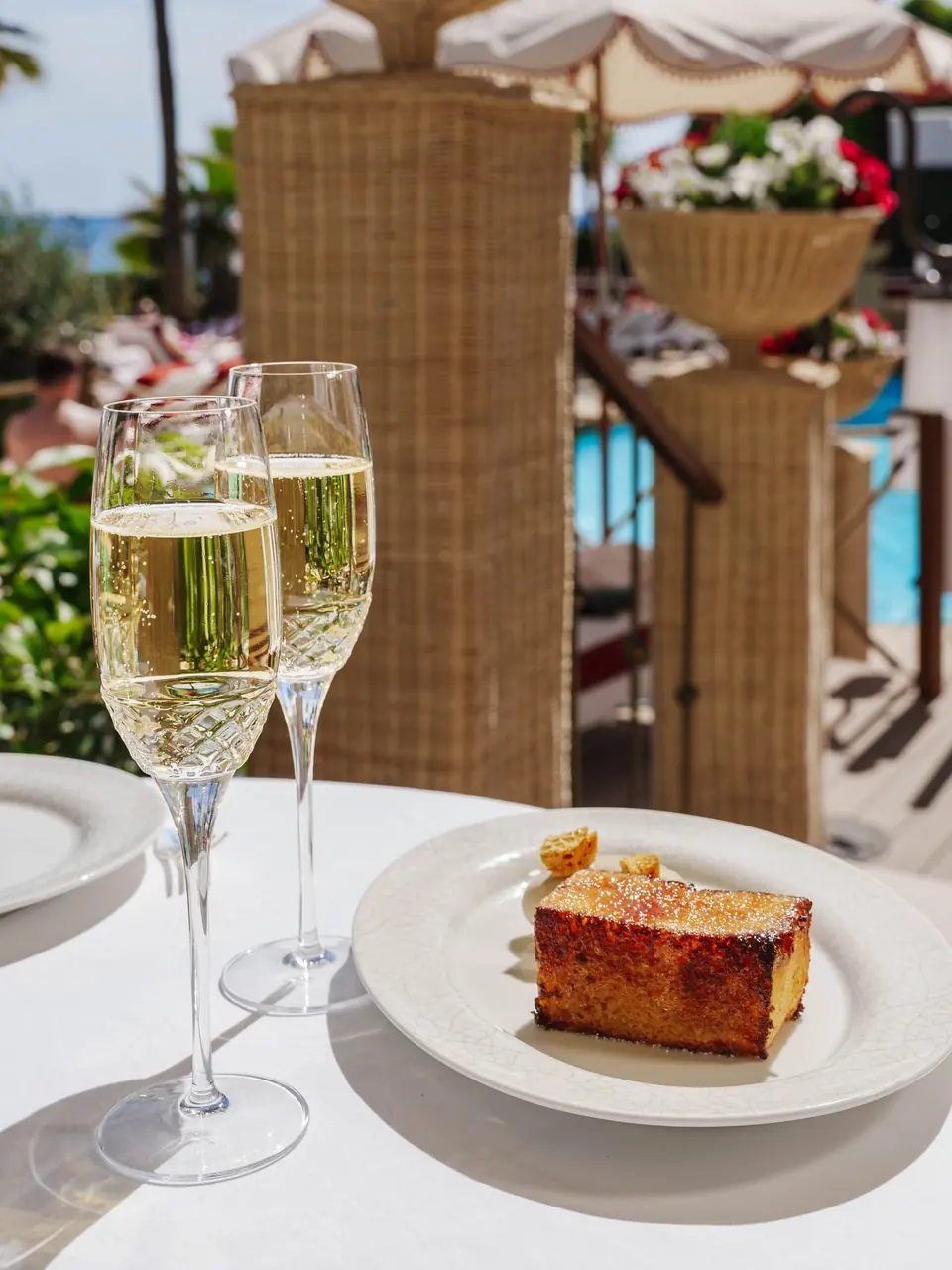 A slice of golden cake beside two glasses of champagne by the pool