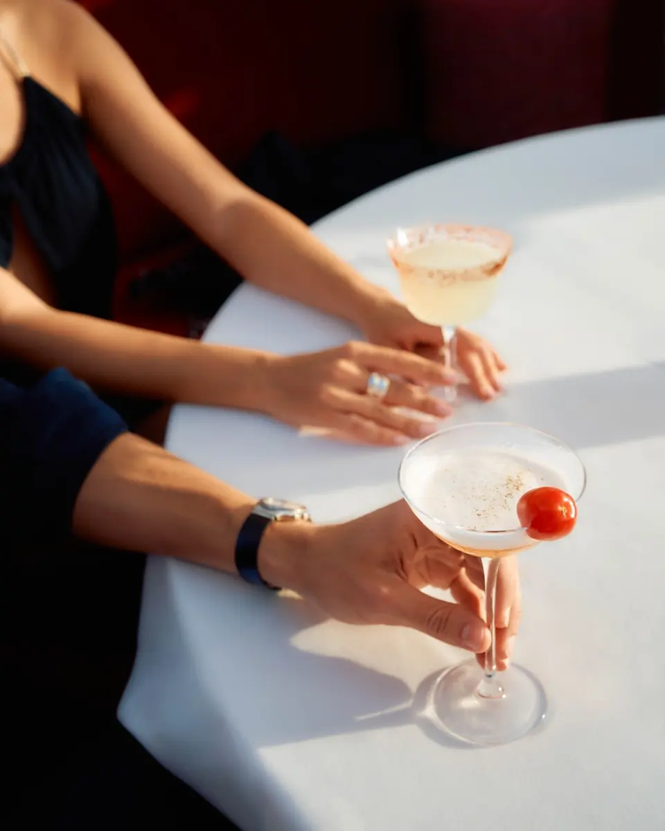 Two hands holding coupe cocktails across a white tablecloth
