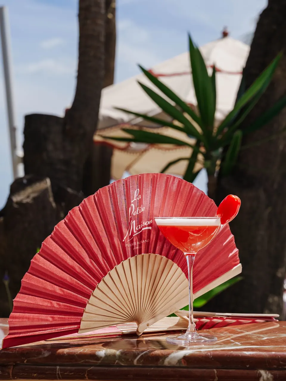 A pink cocktail with a cherry beside an open LPM fan on marble
