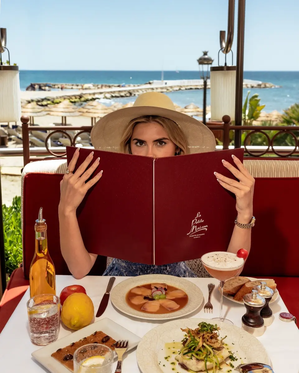 A guest in a sun hat reading the red La Petite Maison menu on the terrace