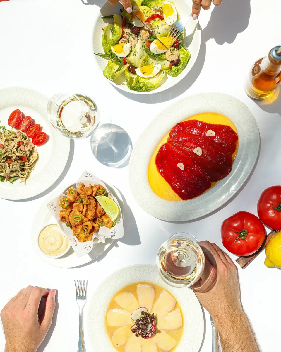A white table seen from above, set with Salade Niçoise, roasted peppers, fried squid, tomatoes and glasses of rosé