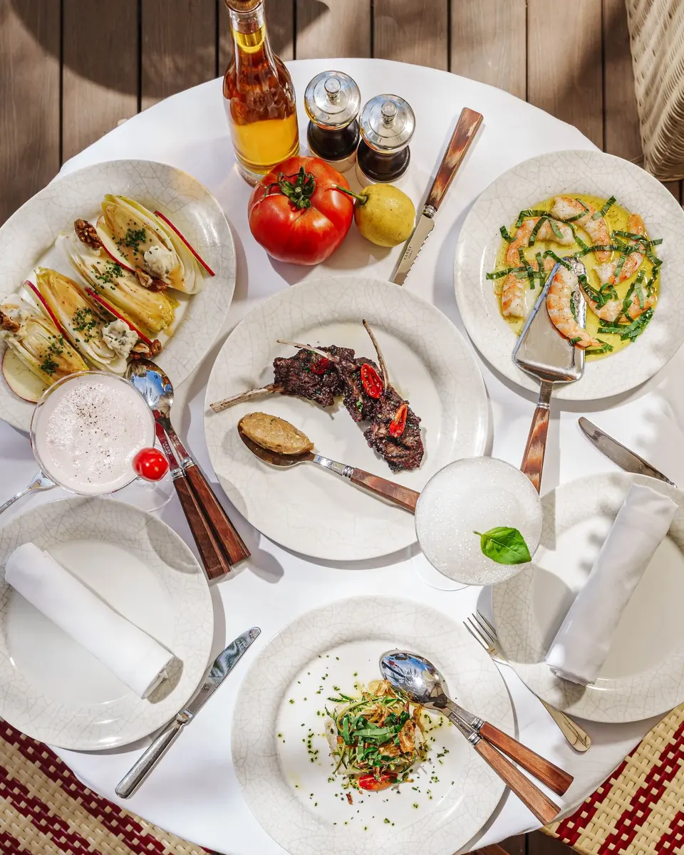 A terrace table seen from above: endive salad, lamb, prawns, olive oil and tomatoes