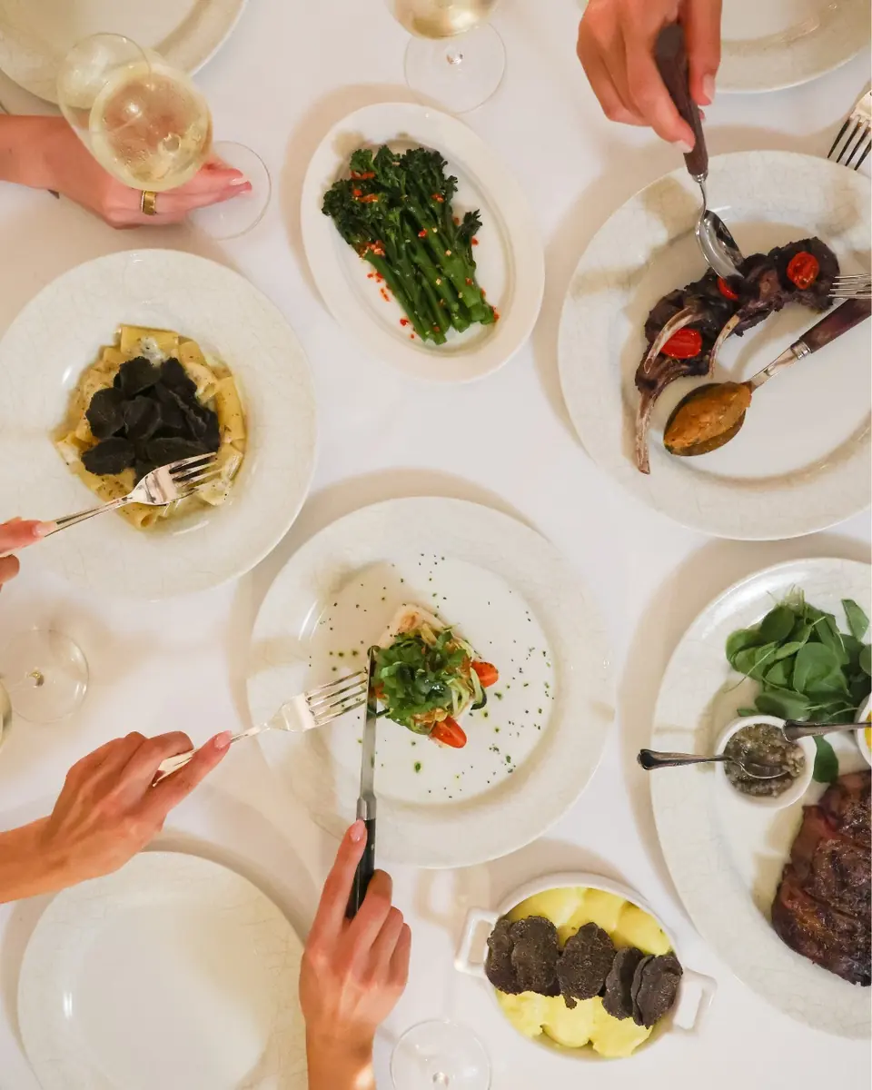 Hands reaching across a shared table of truffle gnocchi, lamb, broccolini and steak