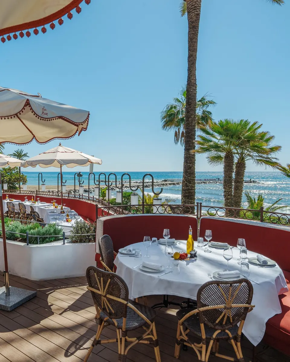 A round table on the terrace under parasols and palms, facing the sea